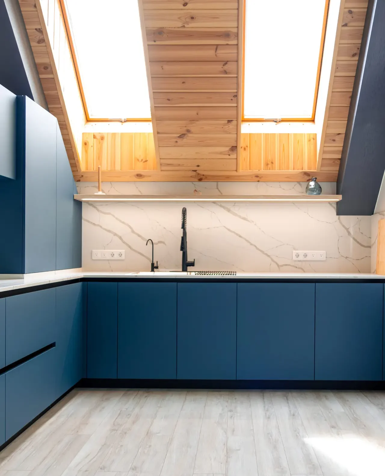 Frontal view of a spacious corner kitchen under skylights with matte blue handleless cabinetry, white marble and quartz. Daylight and LED accents create a soft Scandi vibe.